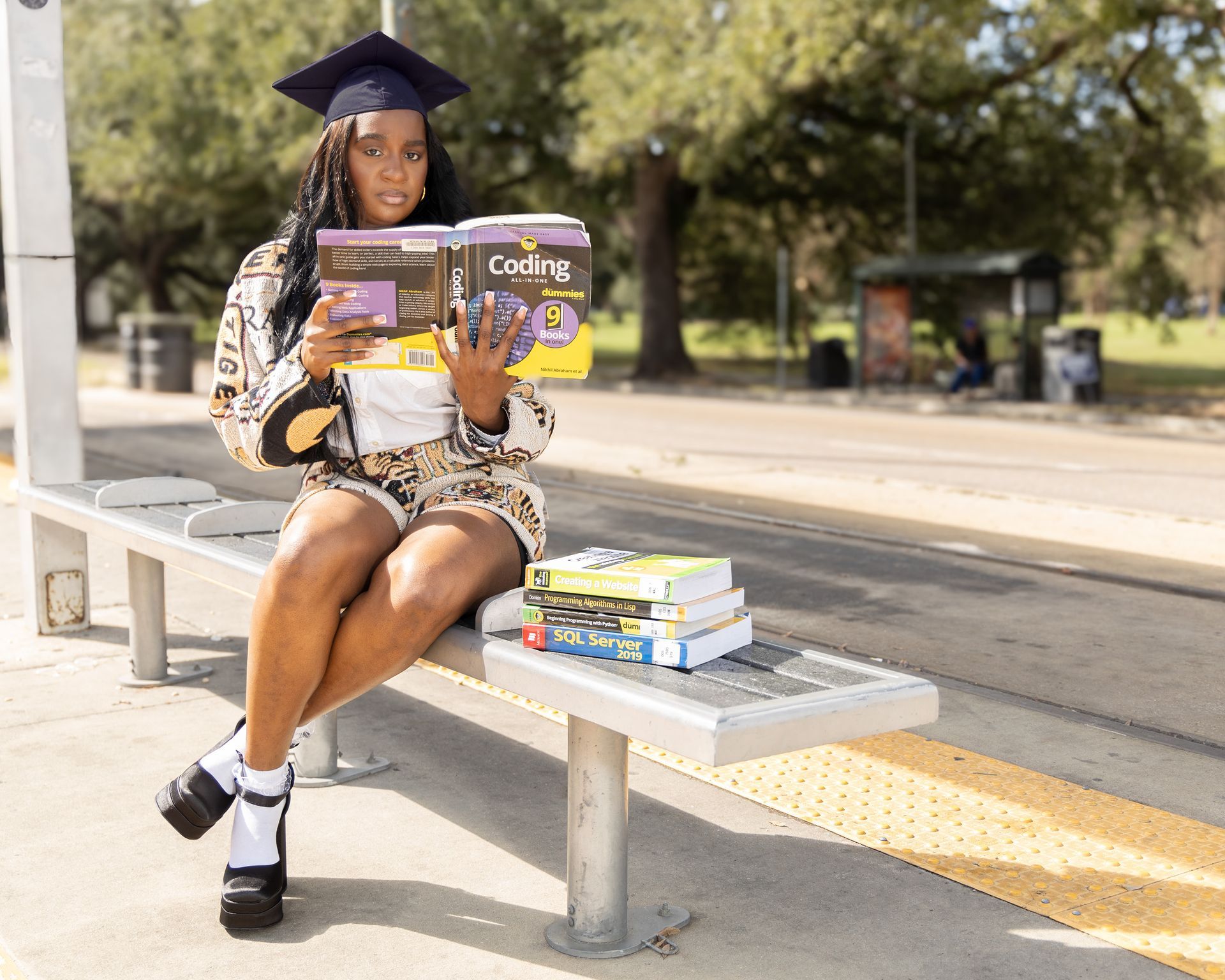 A woman in a graduation cap and gown is sitting on a bench holding a book.