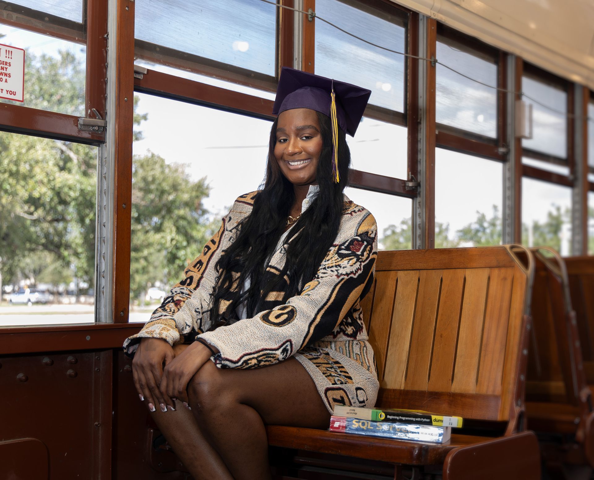 A woman in a graduation cap and gown is sitting on a bus.