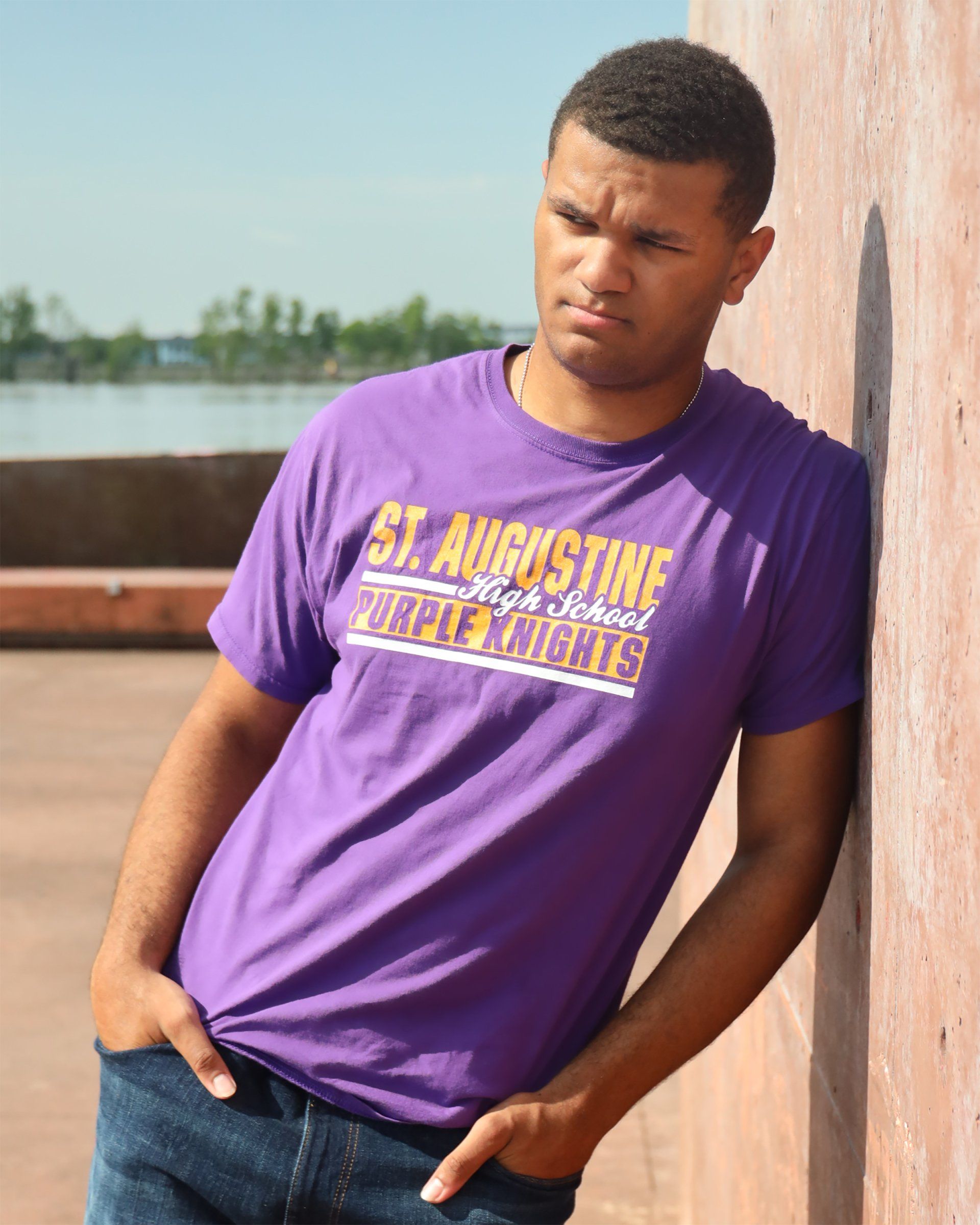 A man leaning against a wall wearing a purple shirt that says st. augustine