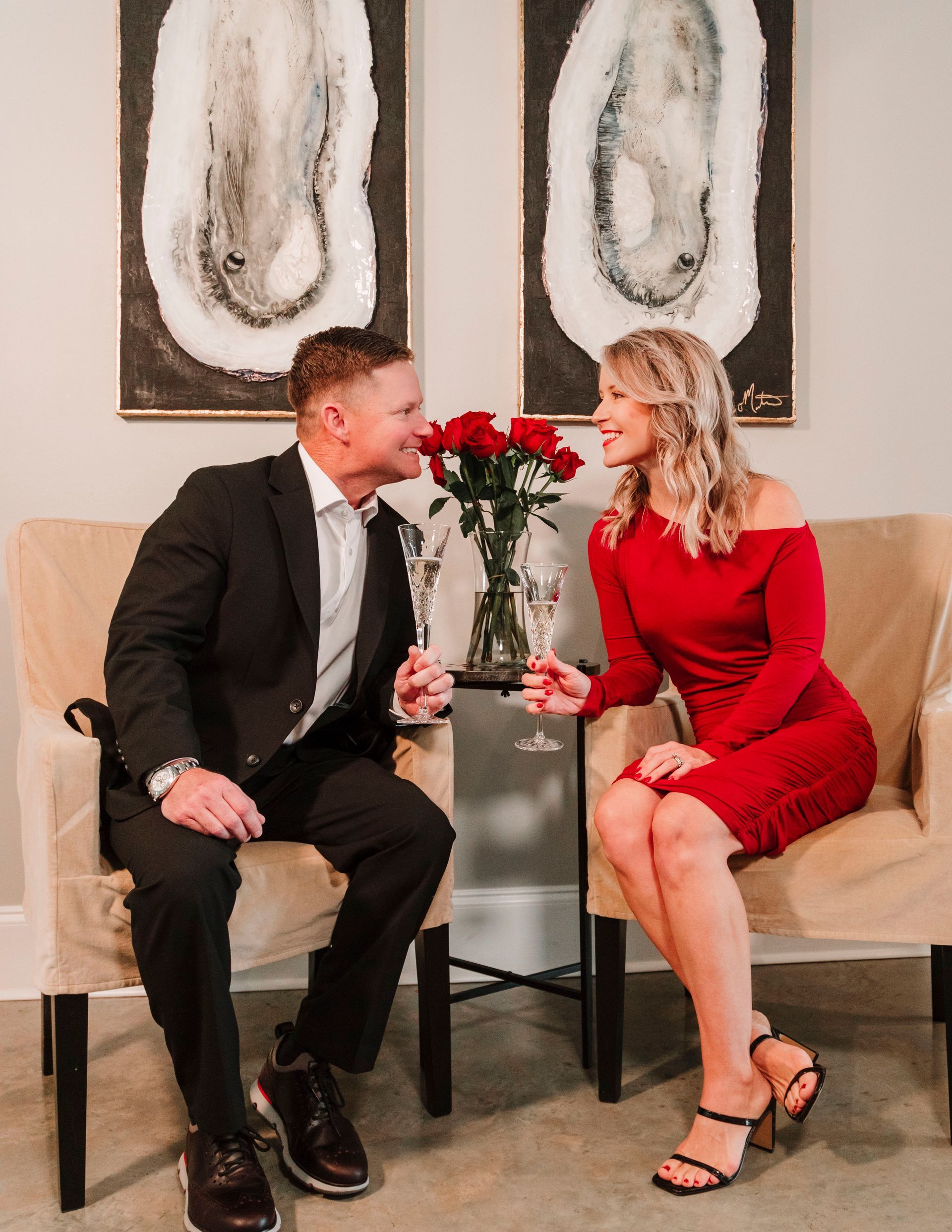 A man in a suit and a woman in a red dress are sitting in chairs holding wine glasses.