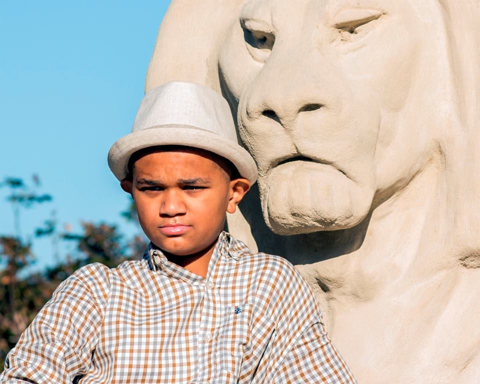 A young boy wearing a hat stands in front of a statue of a lion