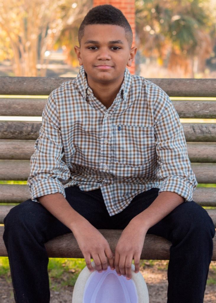 A young boy is sitting on a wooden bench holding a hat.