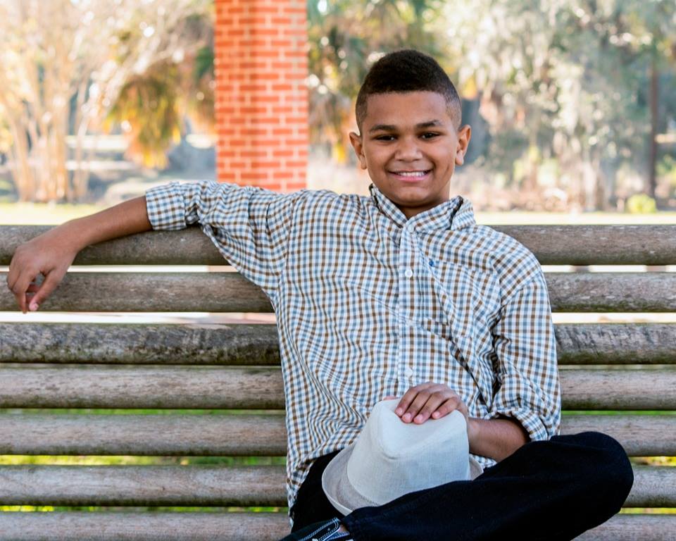 A young boy is sitting on a wooden bench holding a cowboy hat.