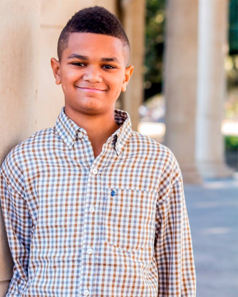 A young boy in a plaid shirt is leaning against a wall and smiling.
