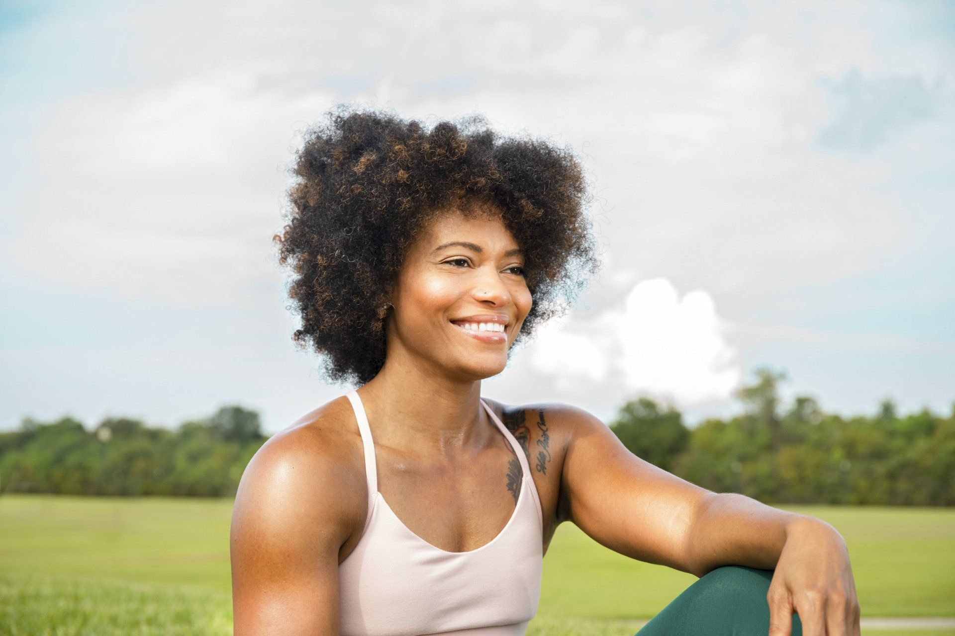 A woman is sitting on a yoga mat in a park and smiling.