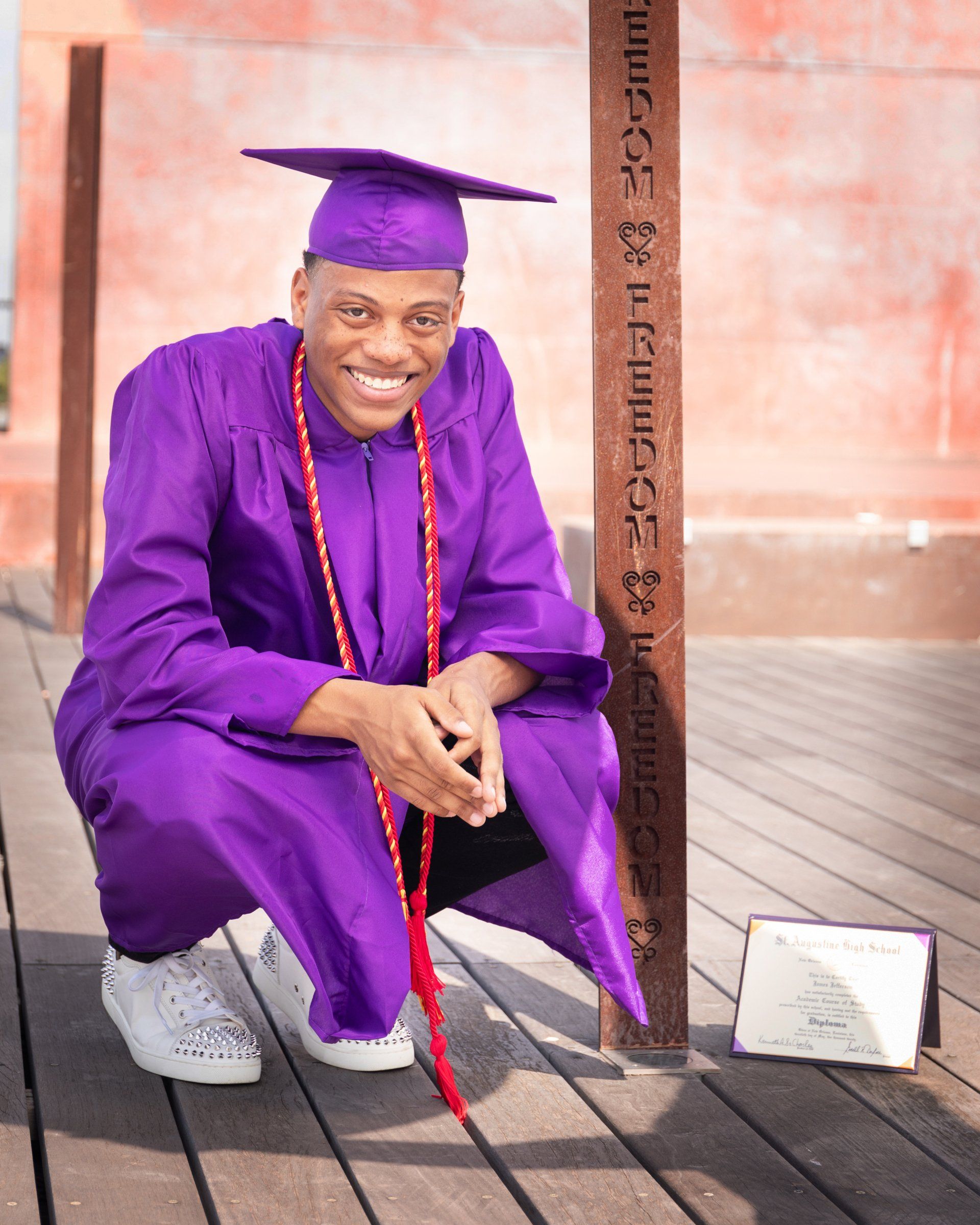 A man in a purple graduation cap and gown is squatting down on a wooden deck.