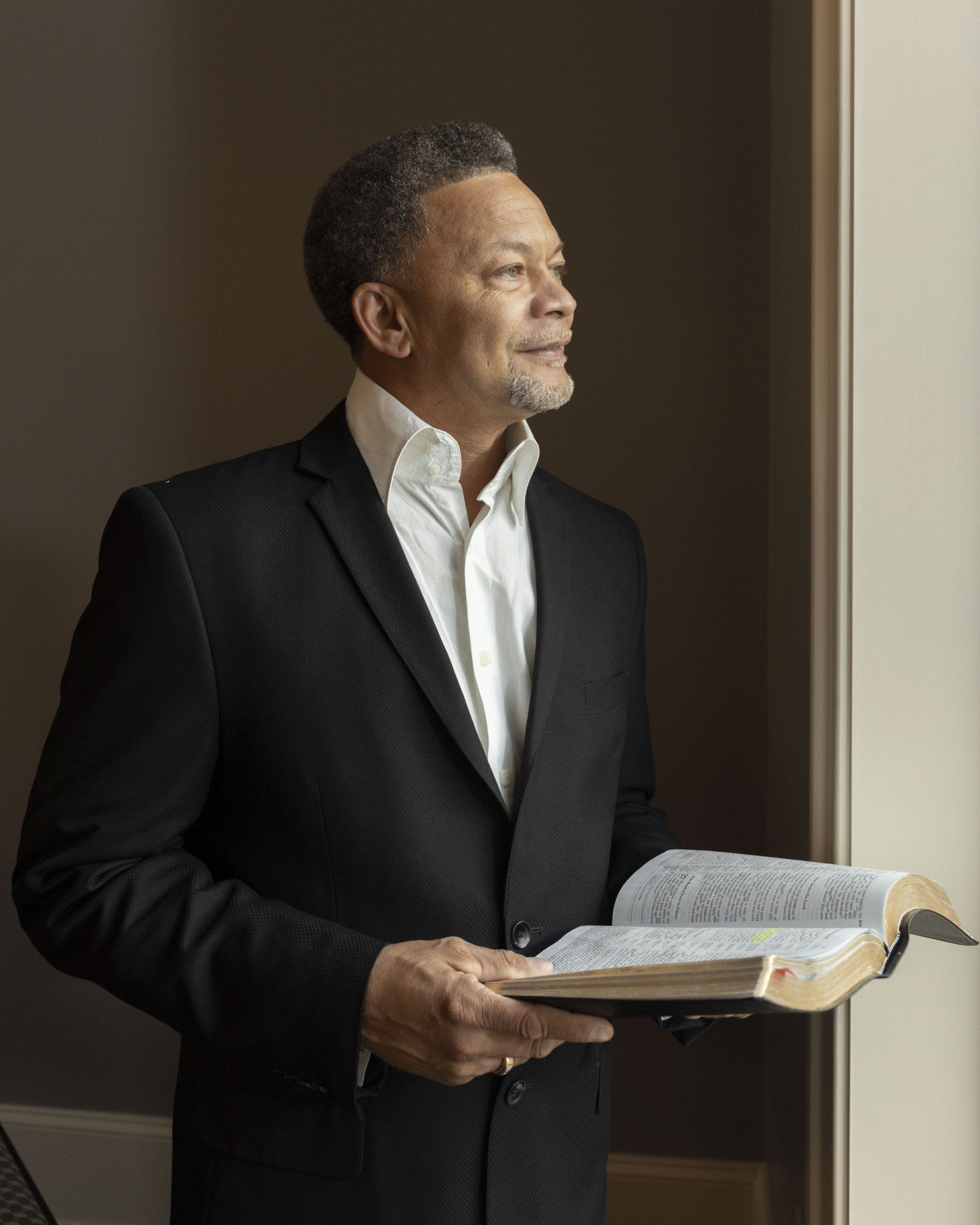 A man in a suit is holding a bible and looking out a window.