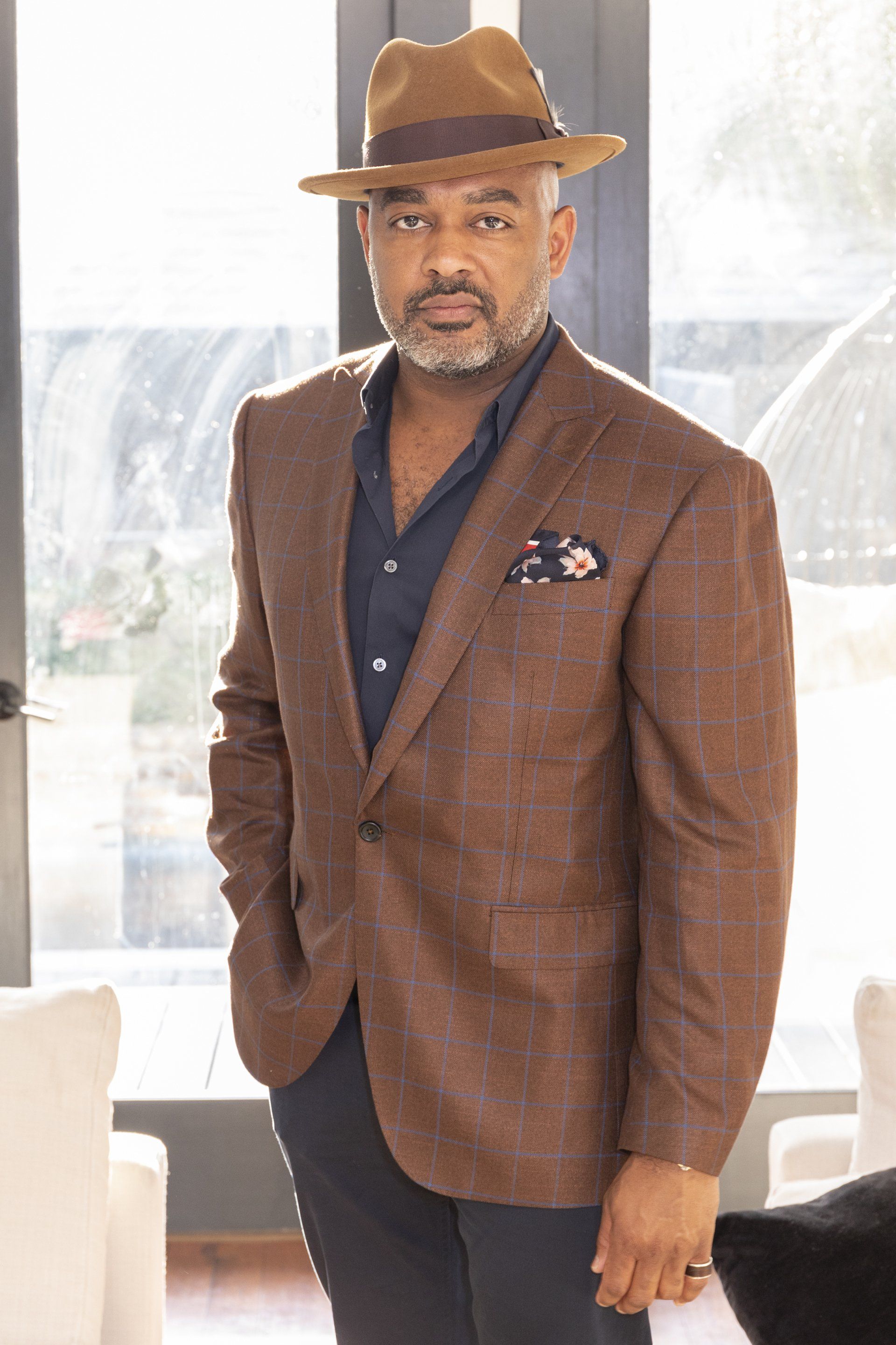 A man wearing a hat and a brown jacket is standing in front of a window.