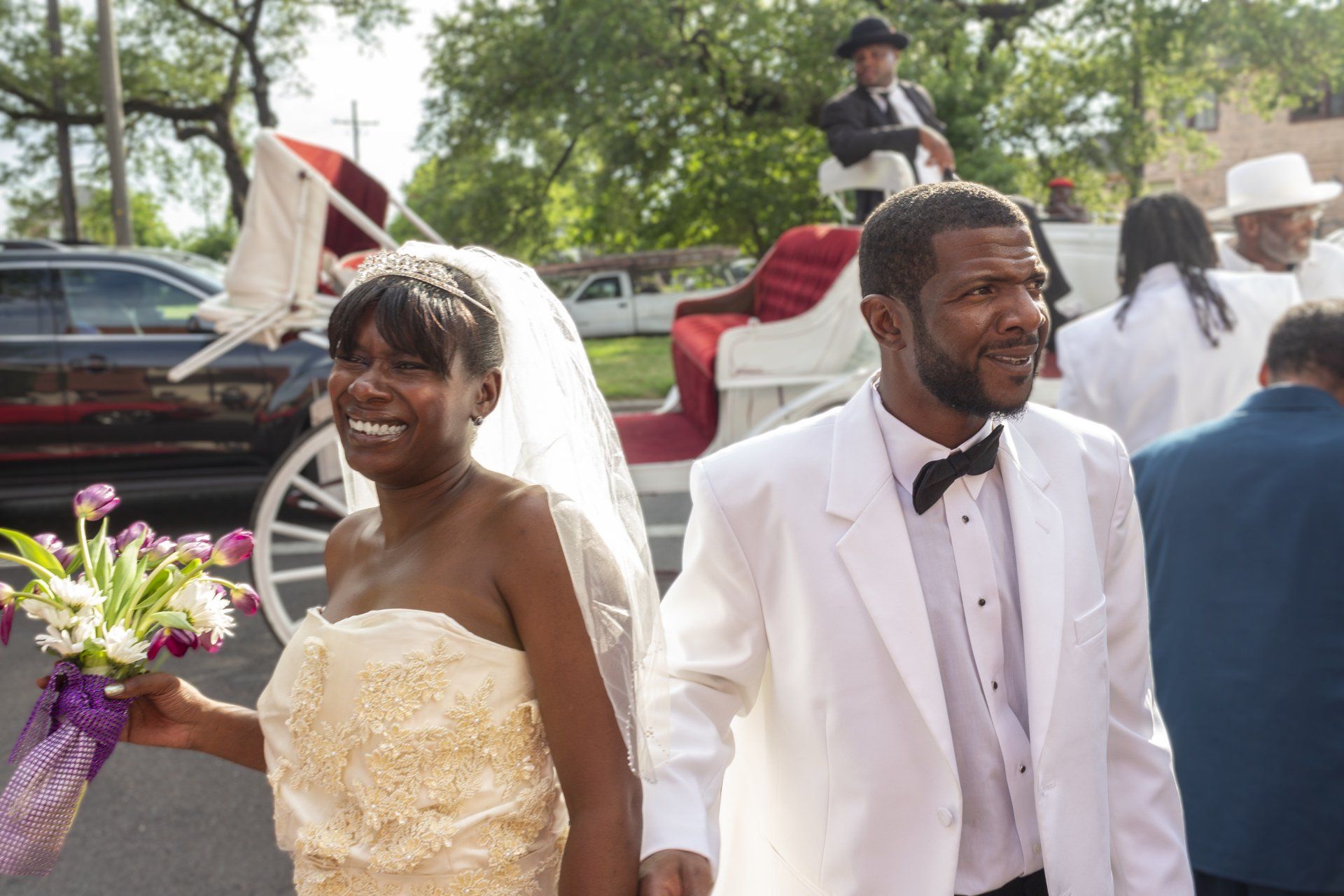 A bride and groom are walking in front of a horse drawn carriage.