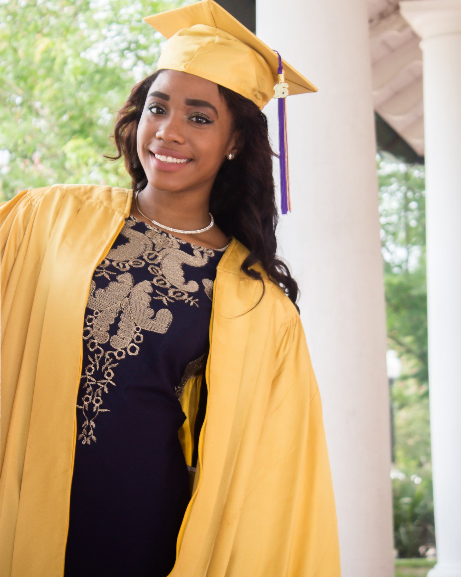 A woman wearing a graduation cap and gown is standing on a porch.