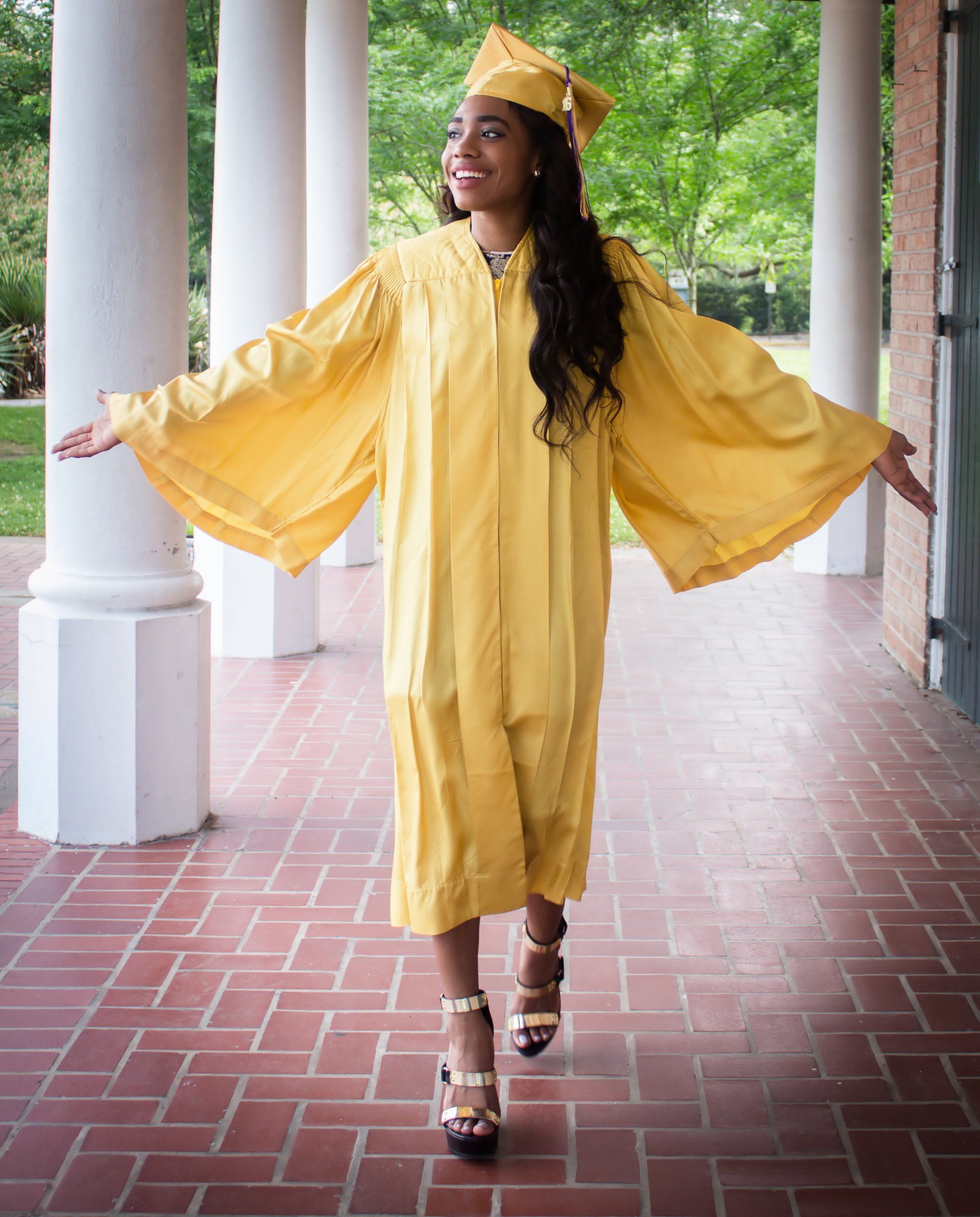 A woman in a yellow graduation cap and gown is standing on a brick walkway.
