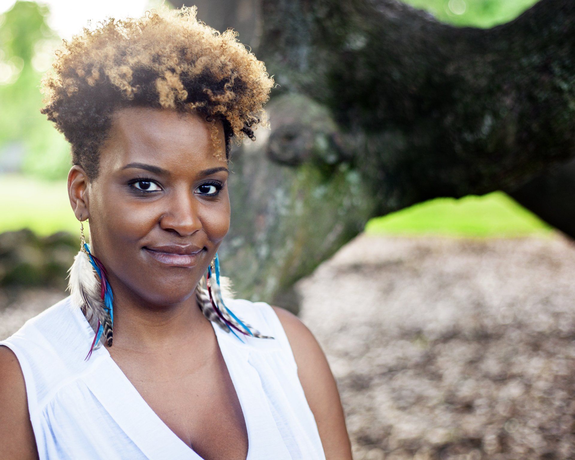 A woman wearing feather earrings and a white shirt is standing in front of a tree.