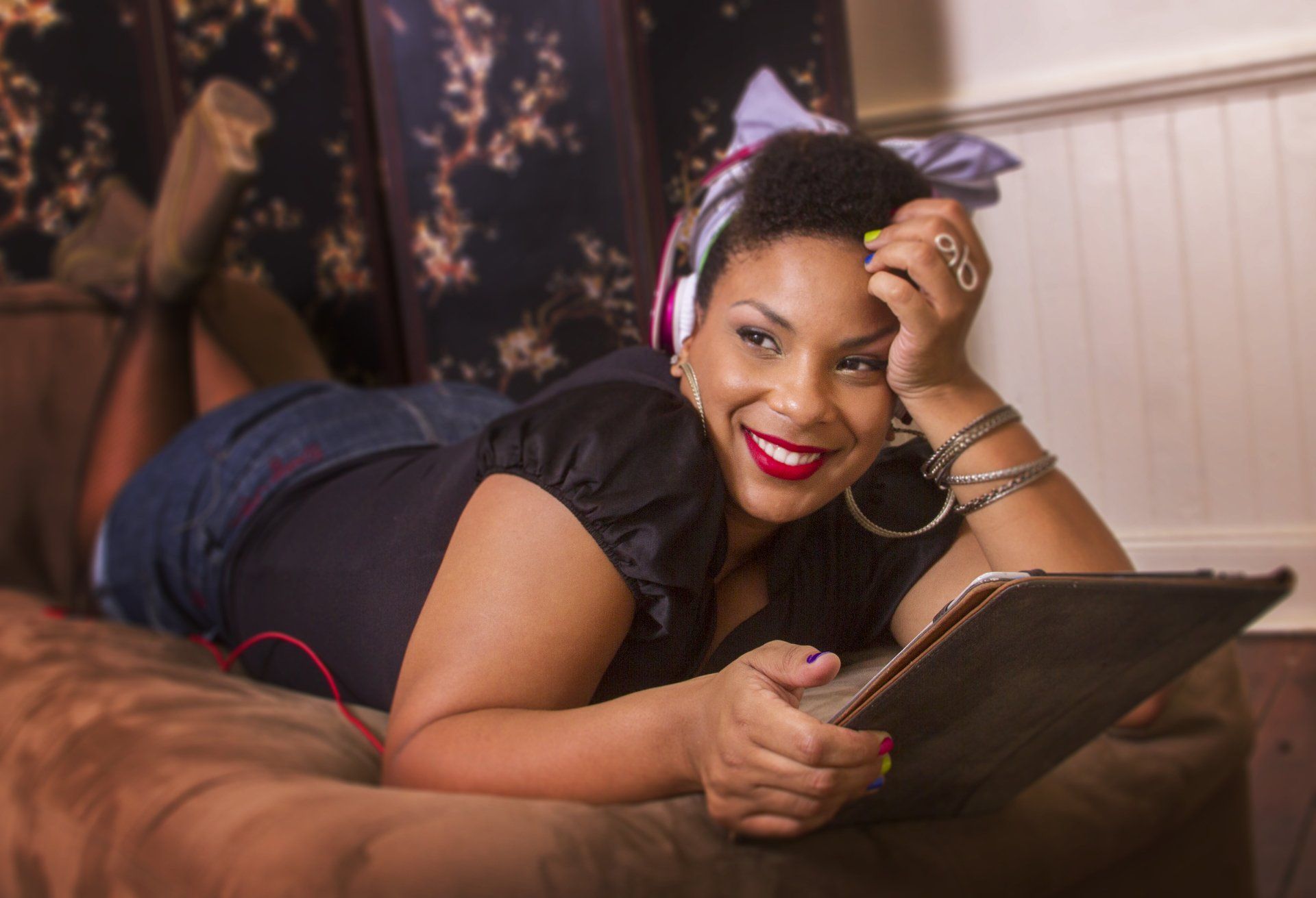 A woman is laying on a couch wearing headphones and holding a tablet.