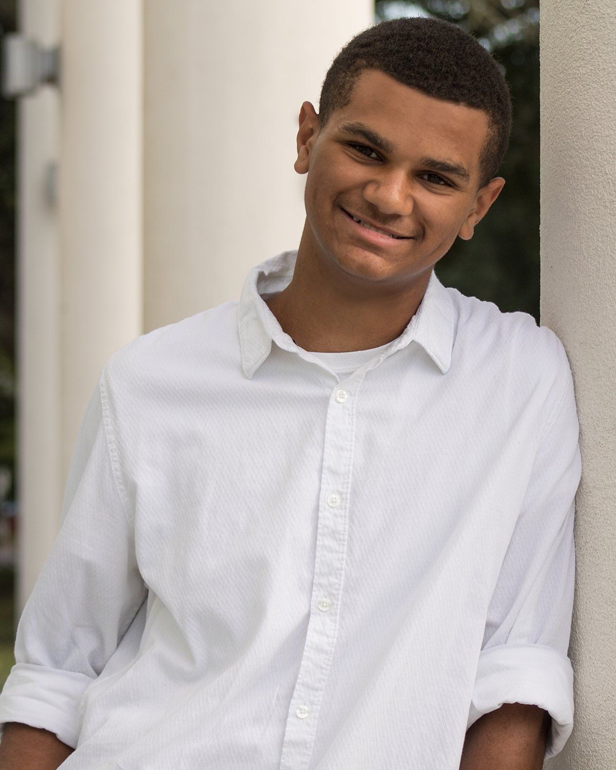 A young man in a white shirt is leaning against a wall and smiling