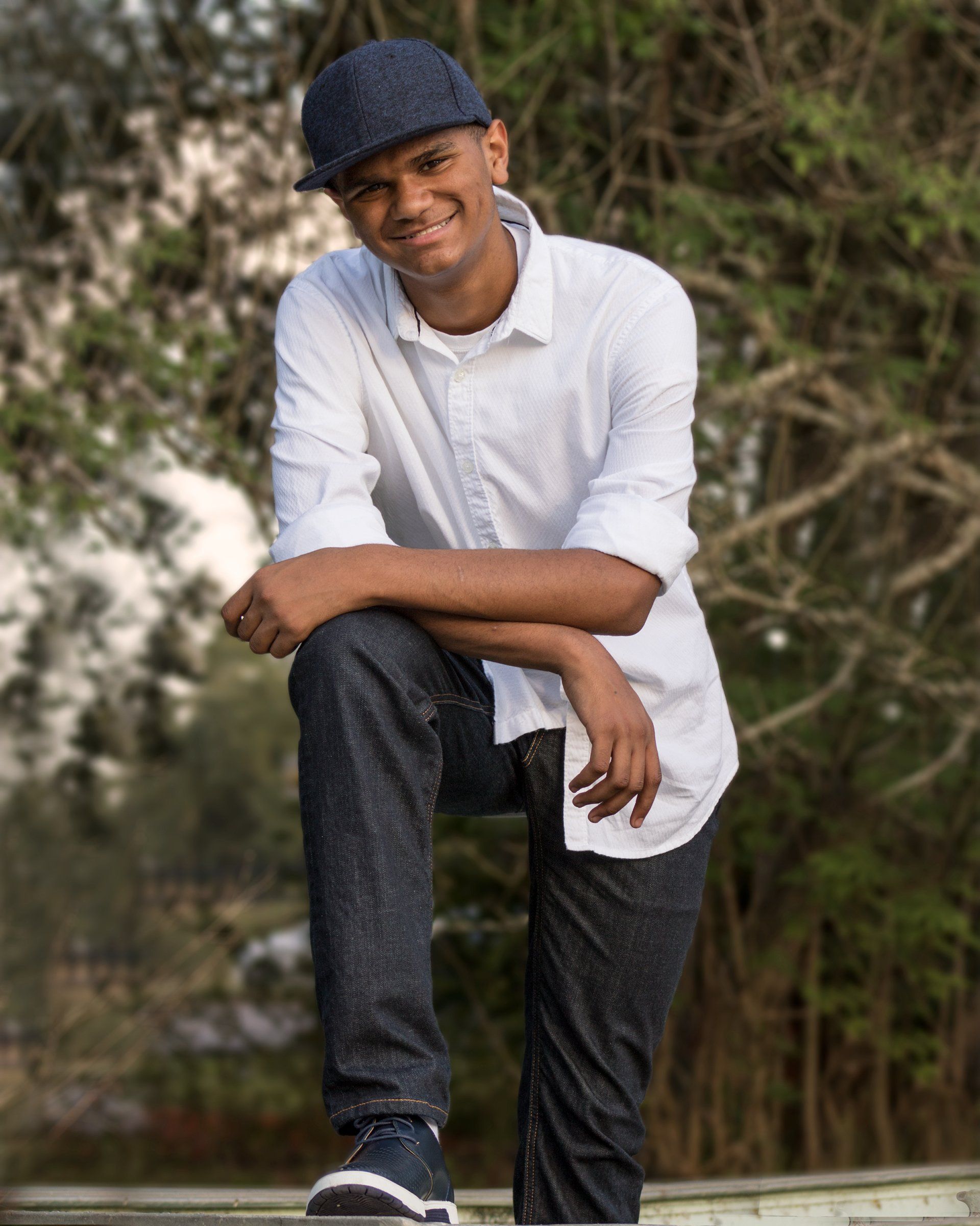 A young man wearing a white shirt and a blue hat is sitting on a railing.