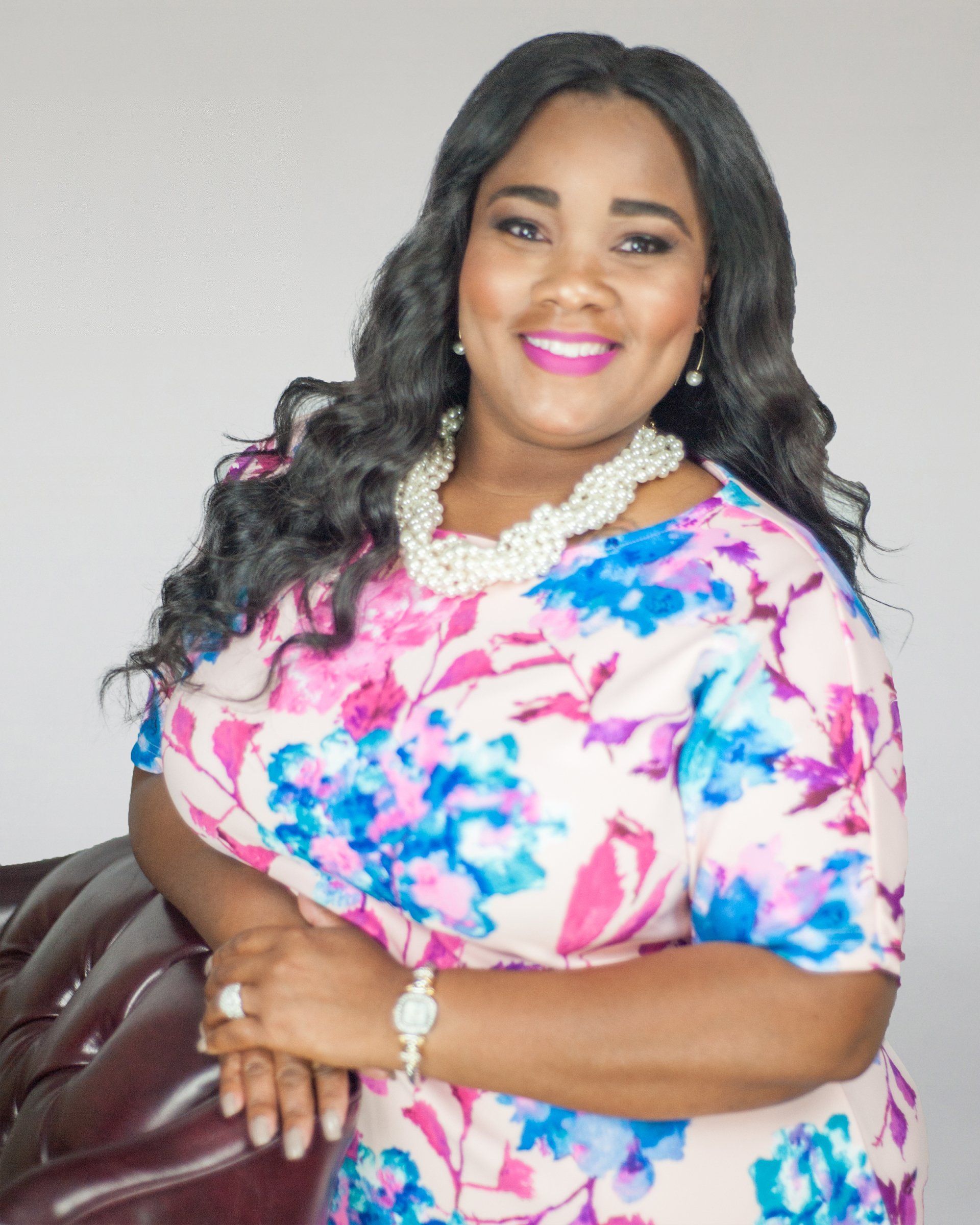 A woman wearing a floral dress and pearls is sitting on a couch.