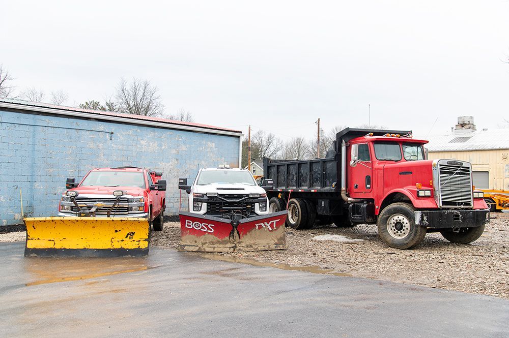 Three work trucks with plows parked outside. A red dump truck stands to the right.