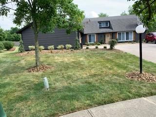 House with dark gray siding and a dormer, on a green lawn with a tree and a lamp post.