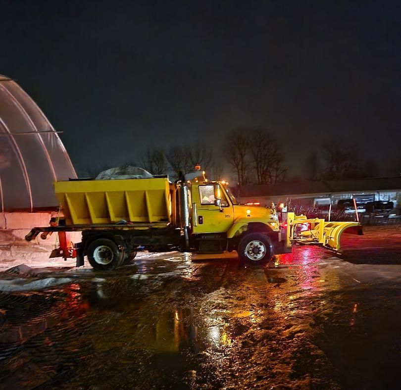 Yellow snowplow truck parked at night, lights on, next to a curved white structure.