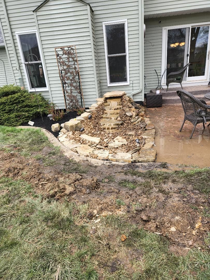 Backyard with a stone waterfall feature, grass, patio, and a house with windows.