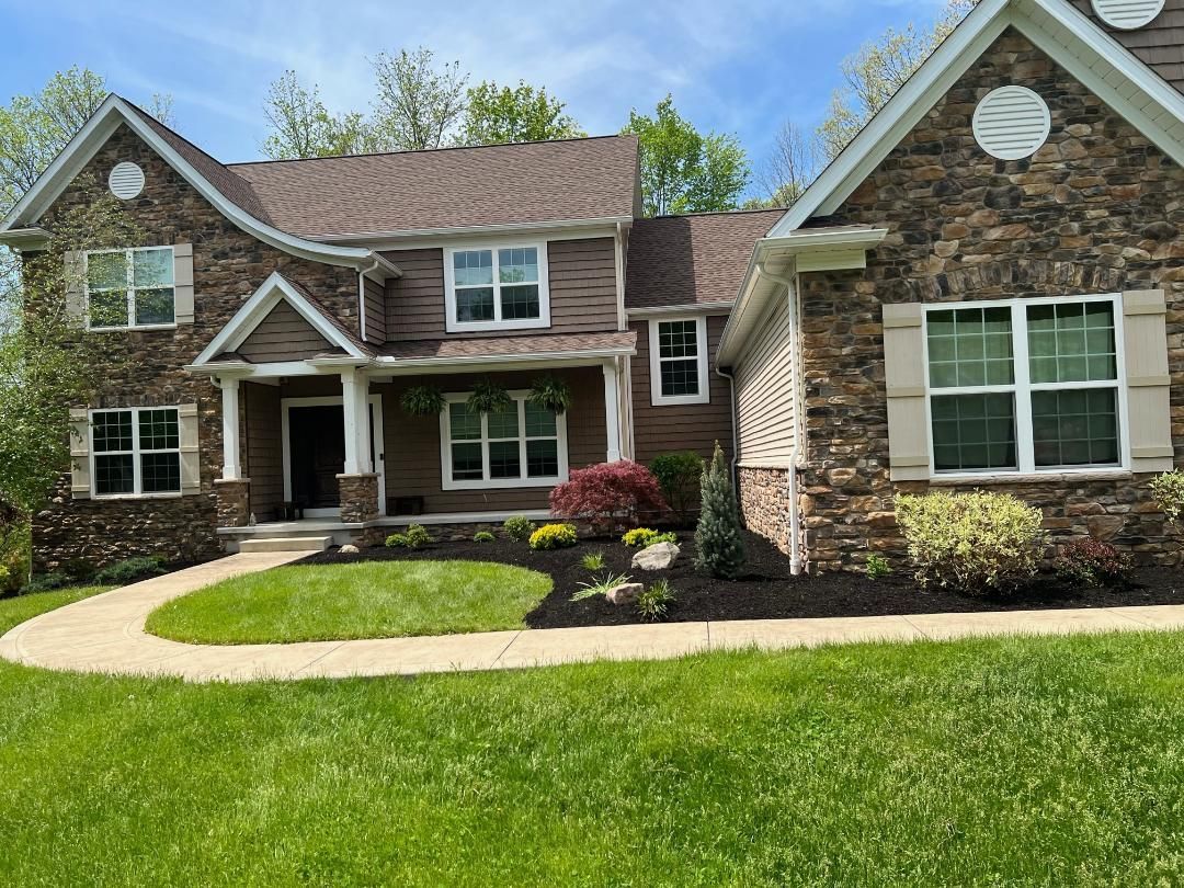 Two-story house with brown and stone exterior, green lawn, and flower bed under a blue sky.