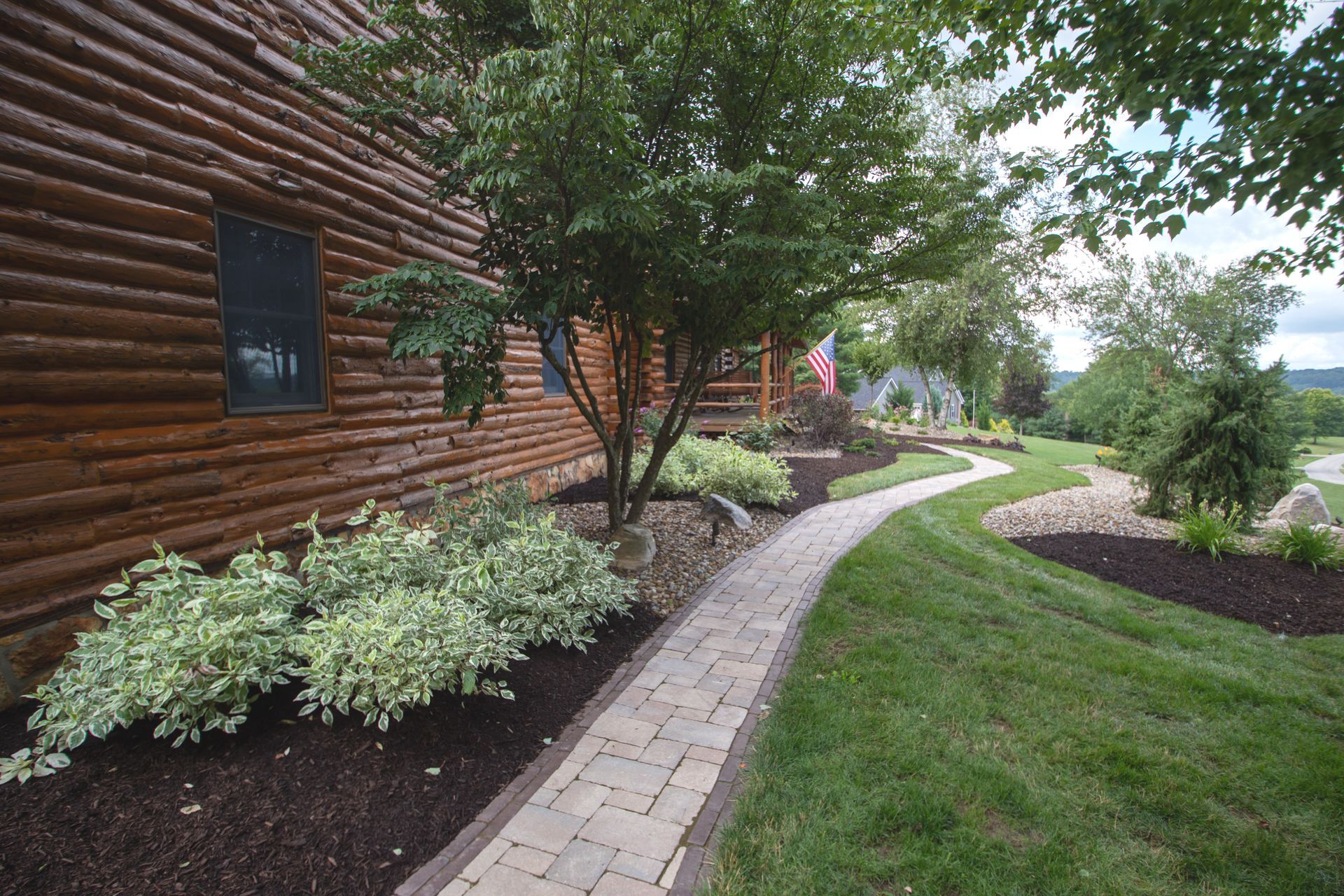 Brick walkway curves through landscaped yard next to a log cabin.