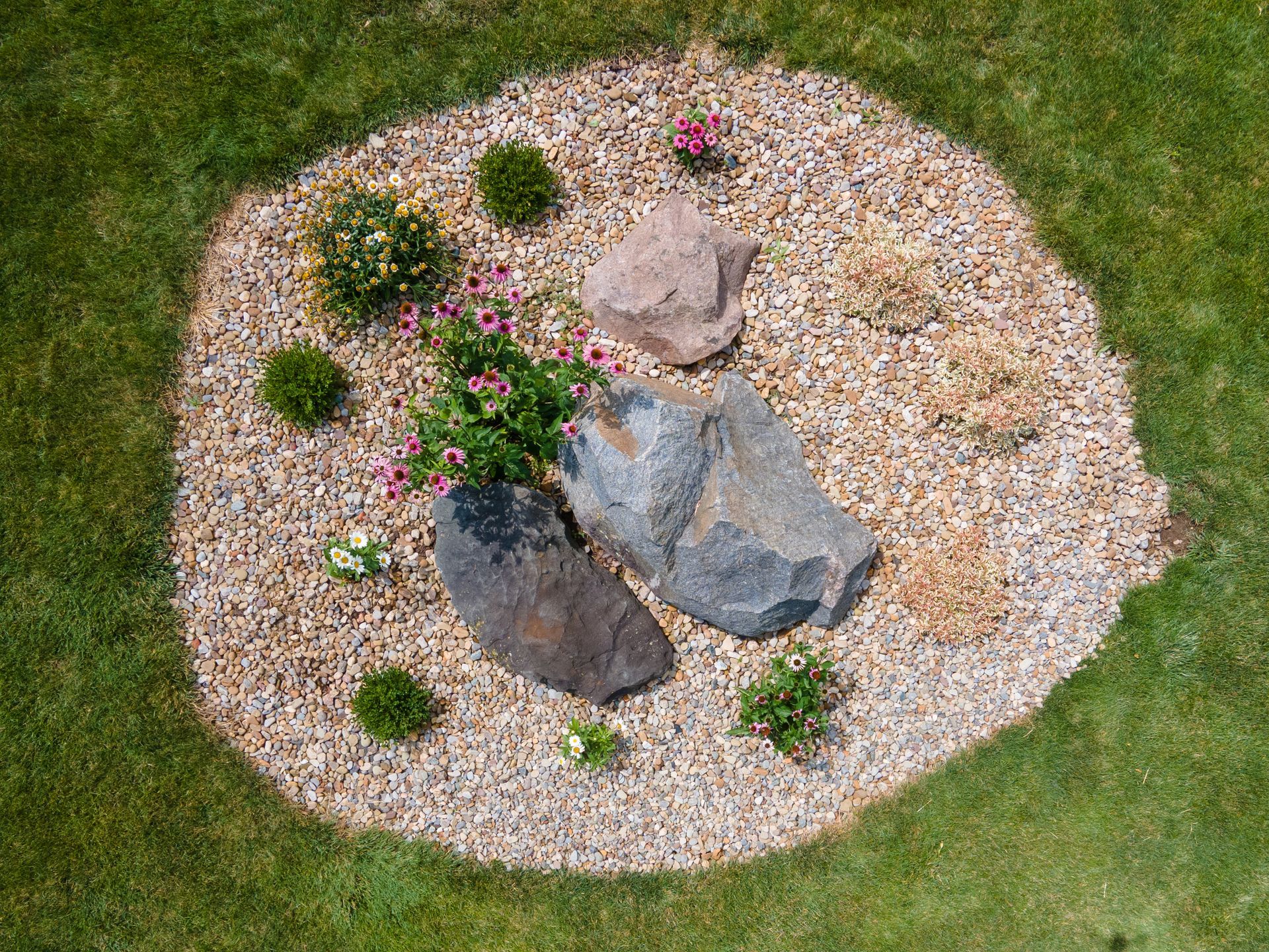 Stone garden bed with rocks, plants, and gravel surrounded by green grass.