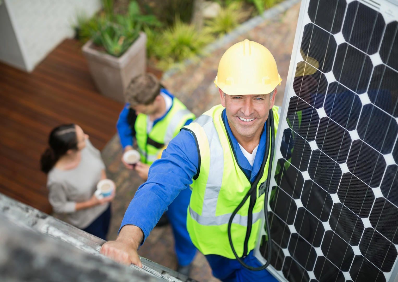 Solar panel installers working on a rooftop, one smiling, holding a panel. Others watching.