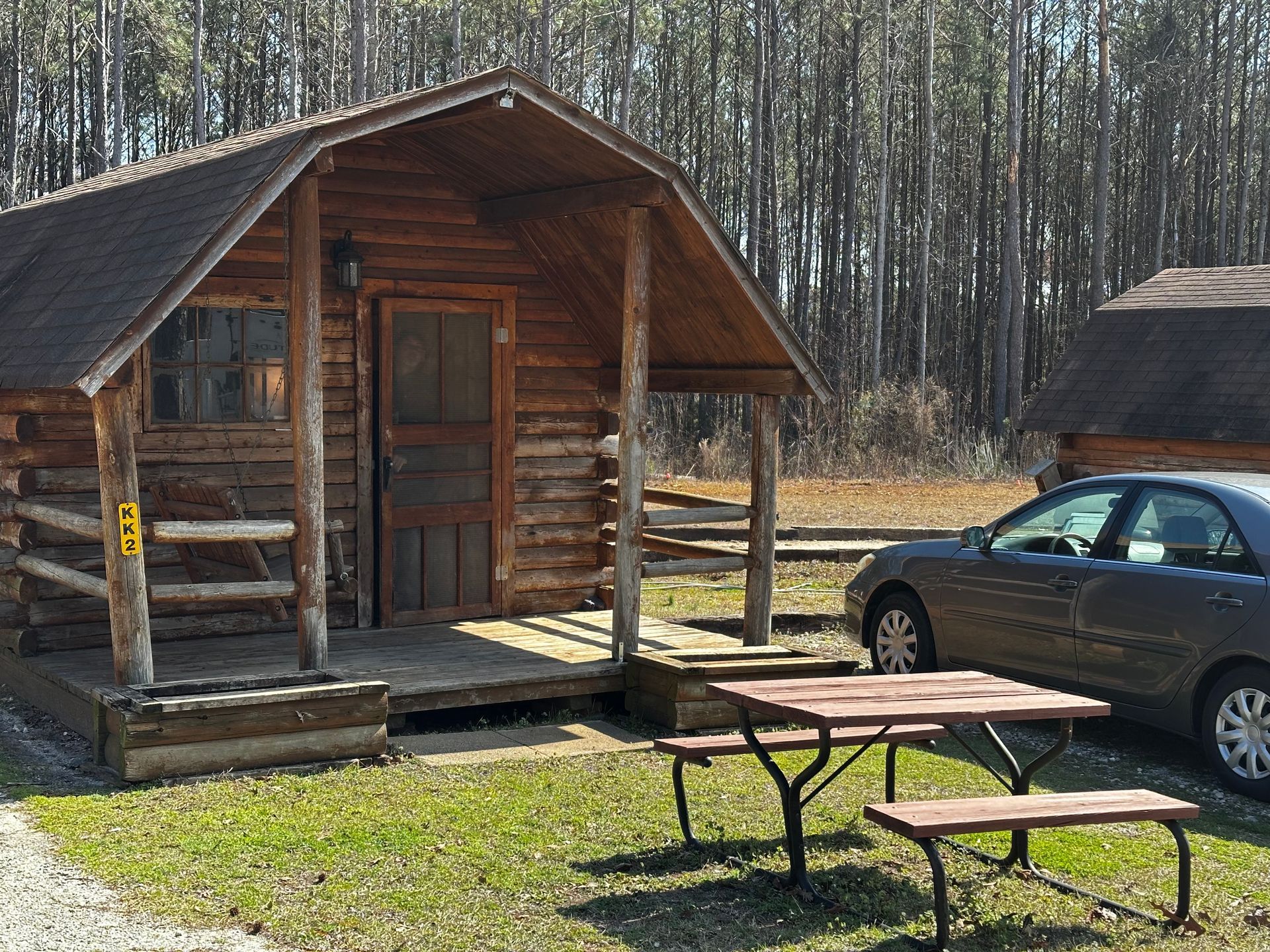 A car is parked in front of a log cabin with a picnic table in front of it.