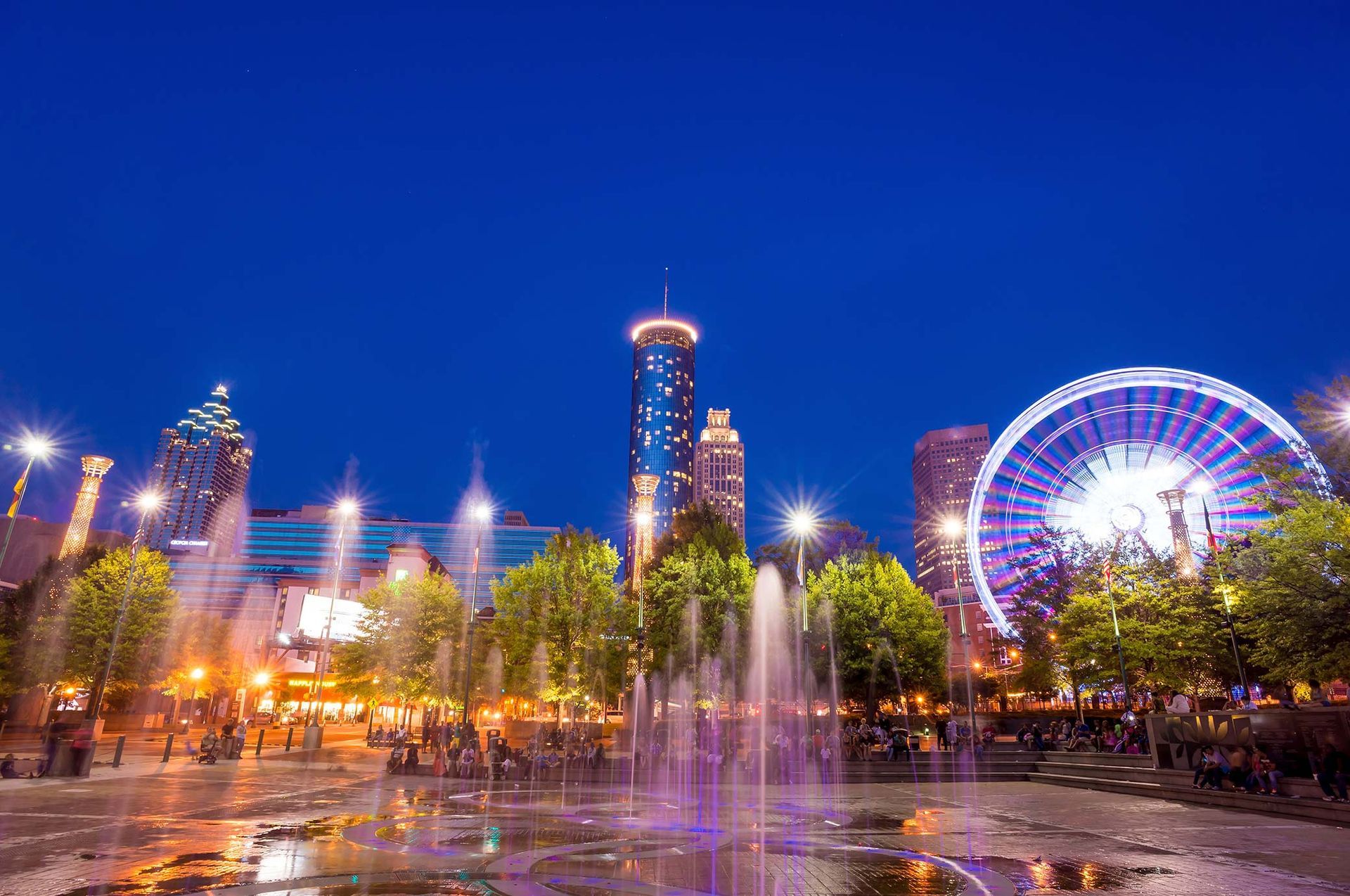 A fountain in a park with a ferris wheel in the background at night.