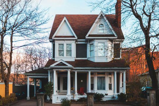 Two-story white house with red roof and porch, framed by bare trees at sunset.