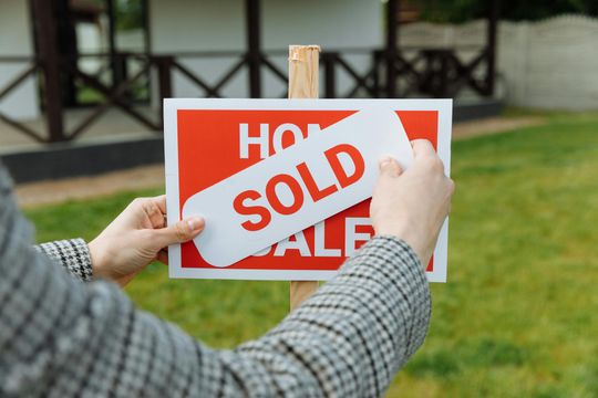 Person holding a red-and-white sold sign with a house in the background