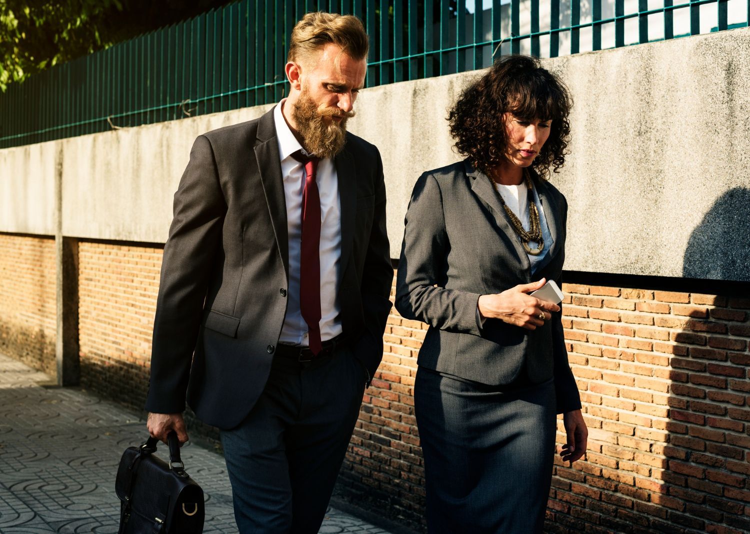 Man with beard and woman in business suits walk side-by-side outdoors.