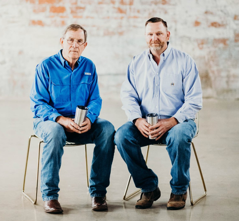 Two men in blue shirts and jeans sit, holding mugs, against a brick wall.