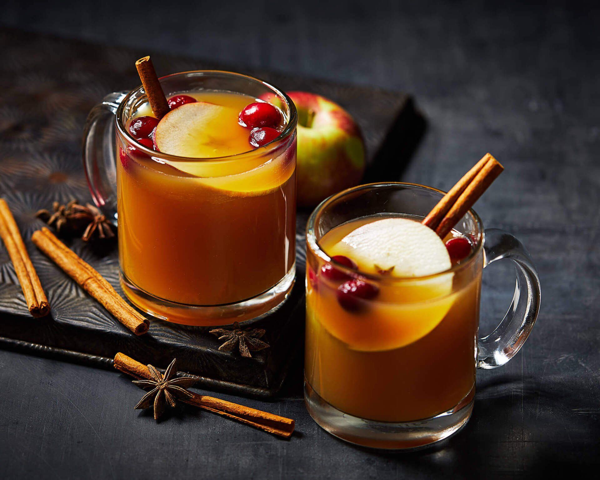 Two cups of apple cider with cinnamon sticks and cranberries on a table.