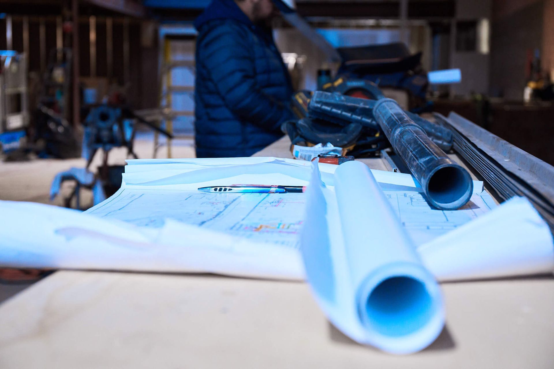 A man is cutting pipes on a table with blueprints and a pen.