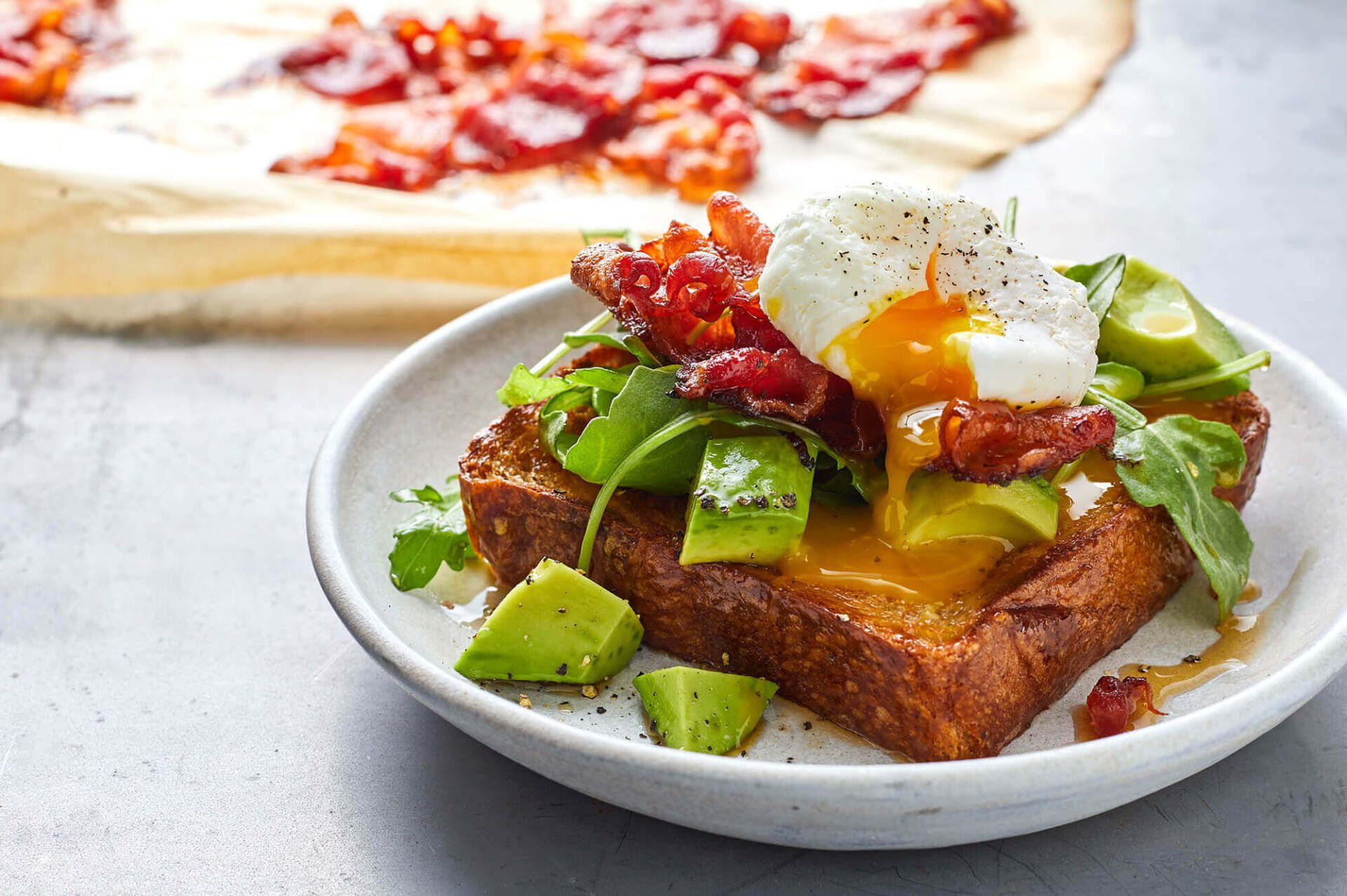 A close up of a plate of food with avocado , bacon and an egg on toast.