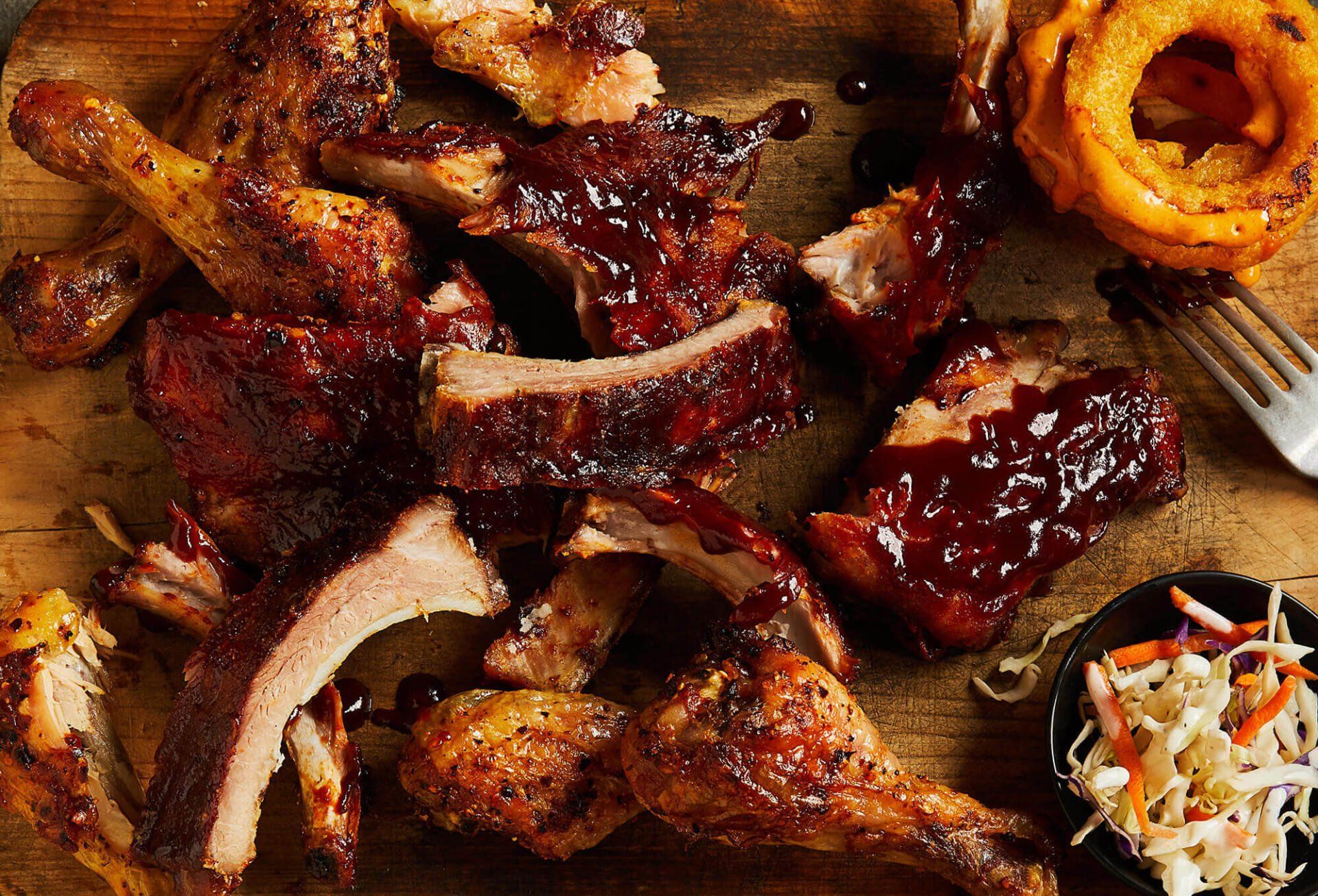A wooden cutting board topped with ribs , chicken , and onion rings.