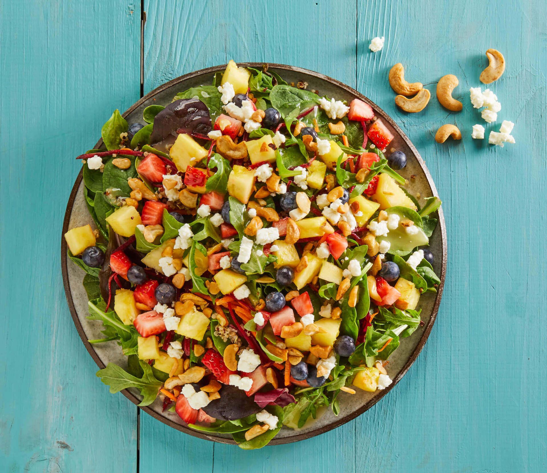 A salad with pineapple , strawberries , blueberries and cashews on a plate on a blue table.