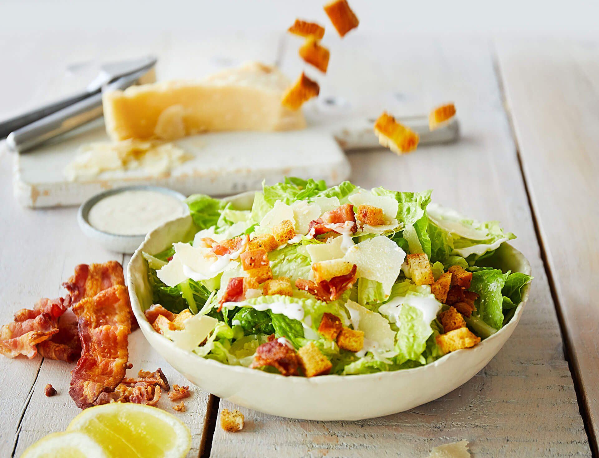 A bowl of caesar salad with croutons and bacon on a table.
