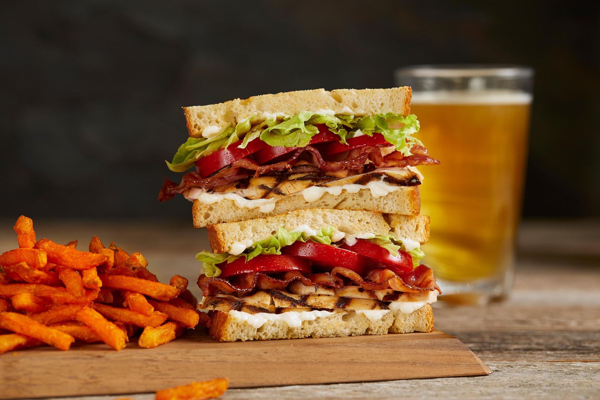 A club sandwich is stacked on top of a cutting board next to french fries and a glass of beer.