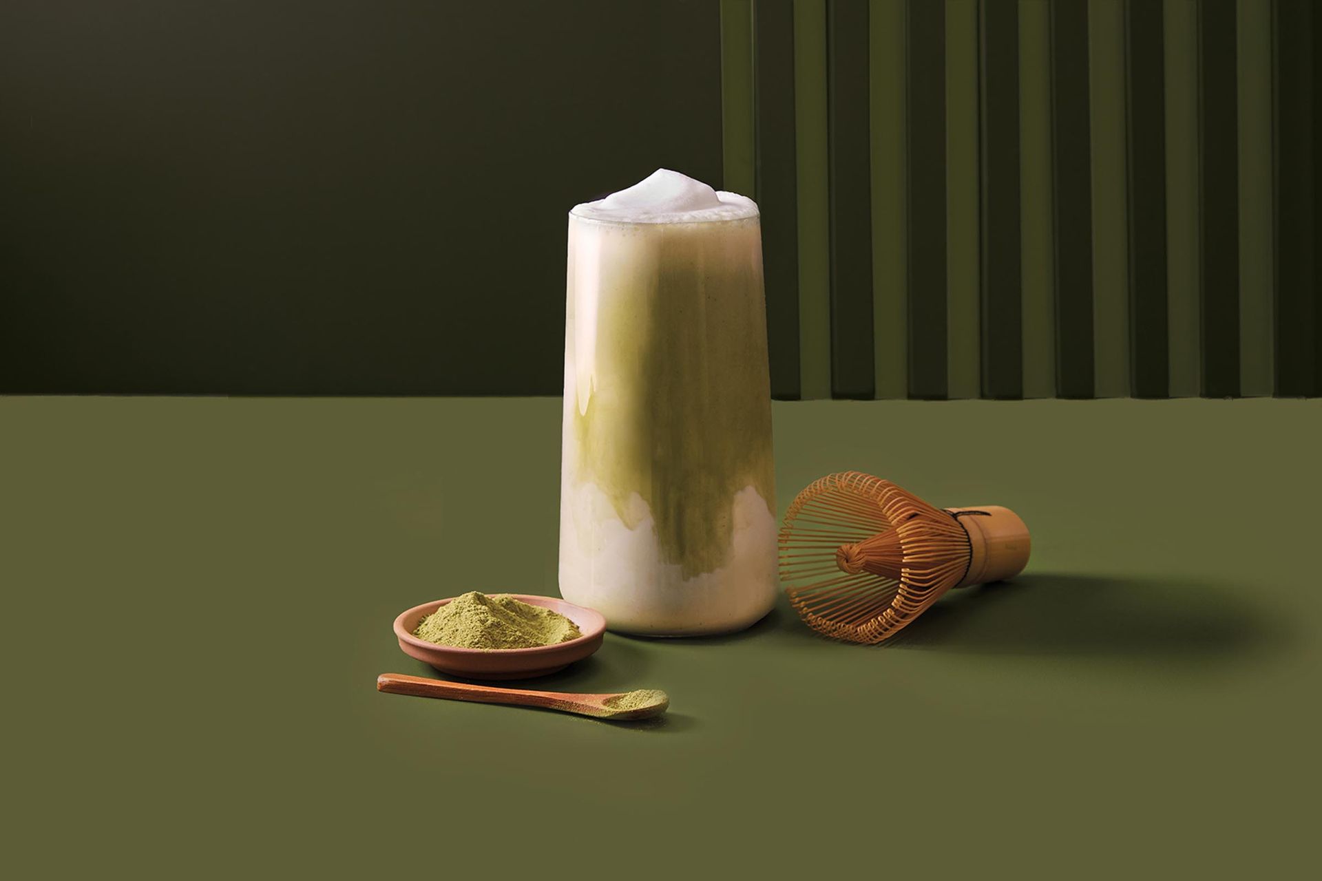 A tall glass filled with a drink next to a bowl of matcha powder and a whisk.