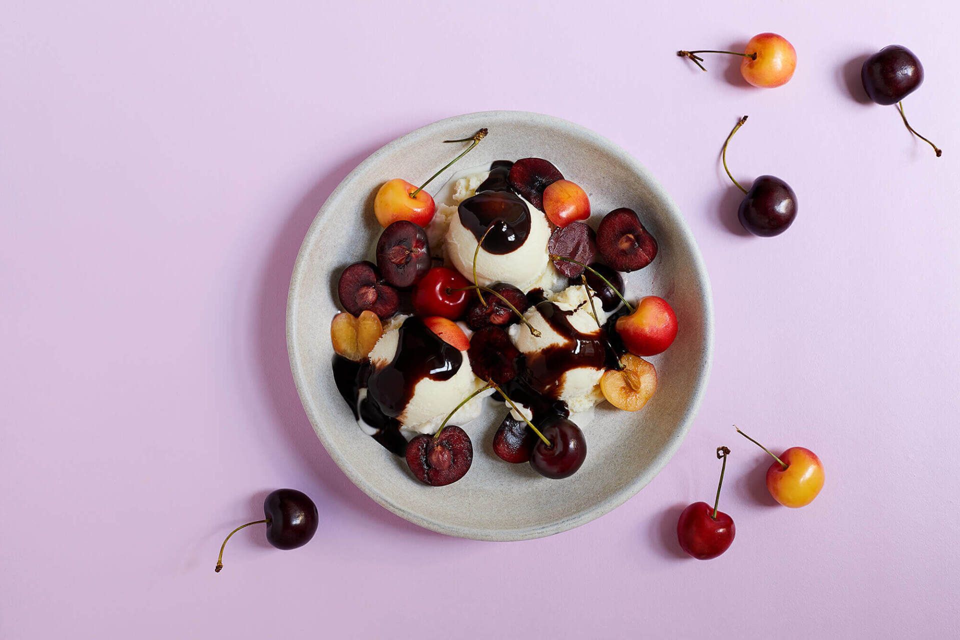 A plate of ice cream with cherries and chocolate sauce on a pink table.