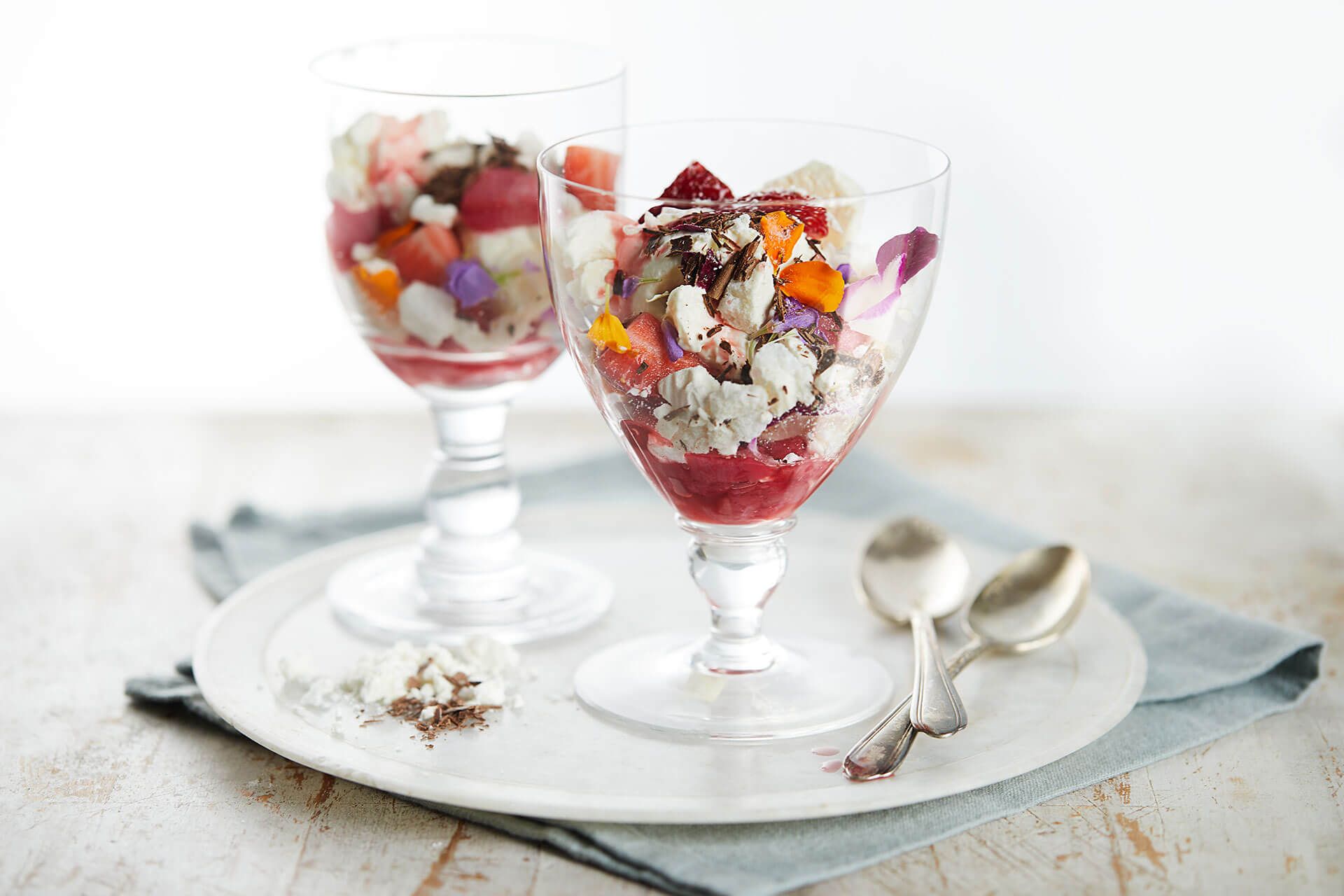 Two desserts in wine glasses on a plate with spoons on a table.