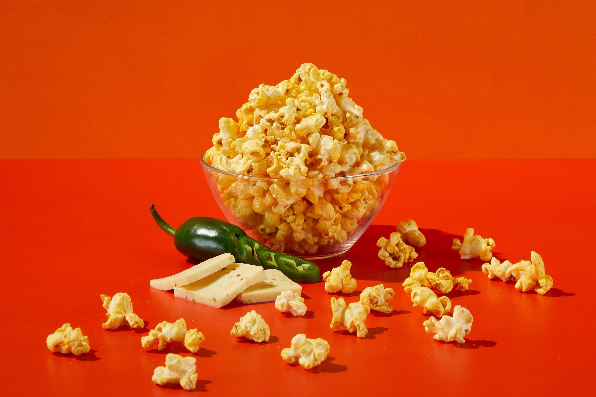 A bowl of popcorn with jalapenos and cheese on an orange table.