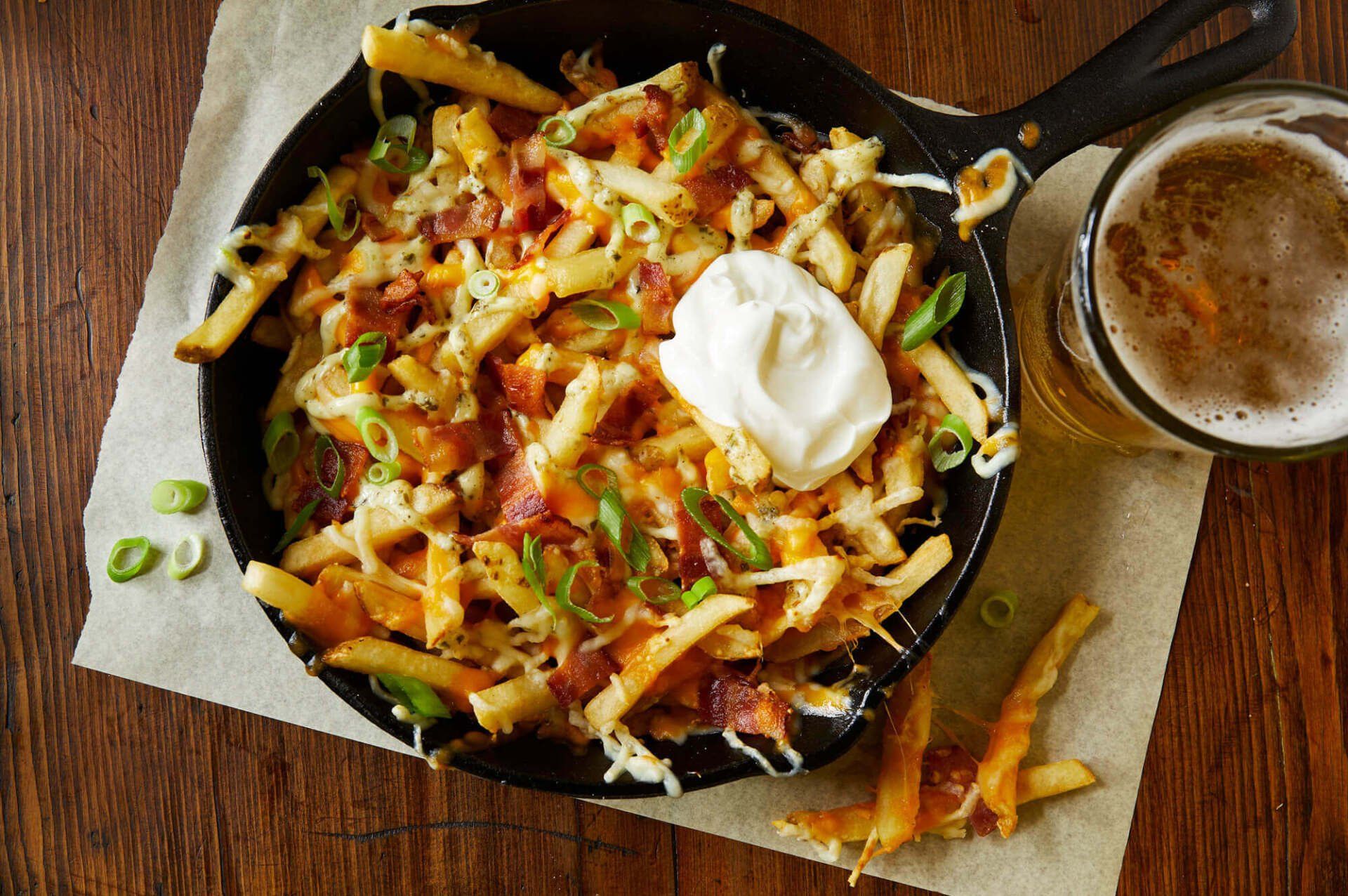A skillet of french fries with sour cream and bacon next to a glass of beer.