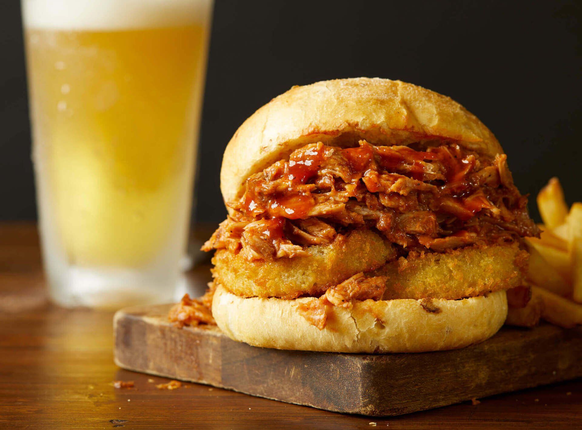 A pulled pork sandwich is sitting on a wooden cutting board next to a glass of beer.
