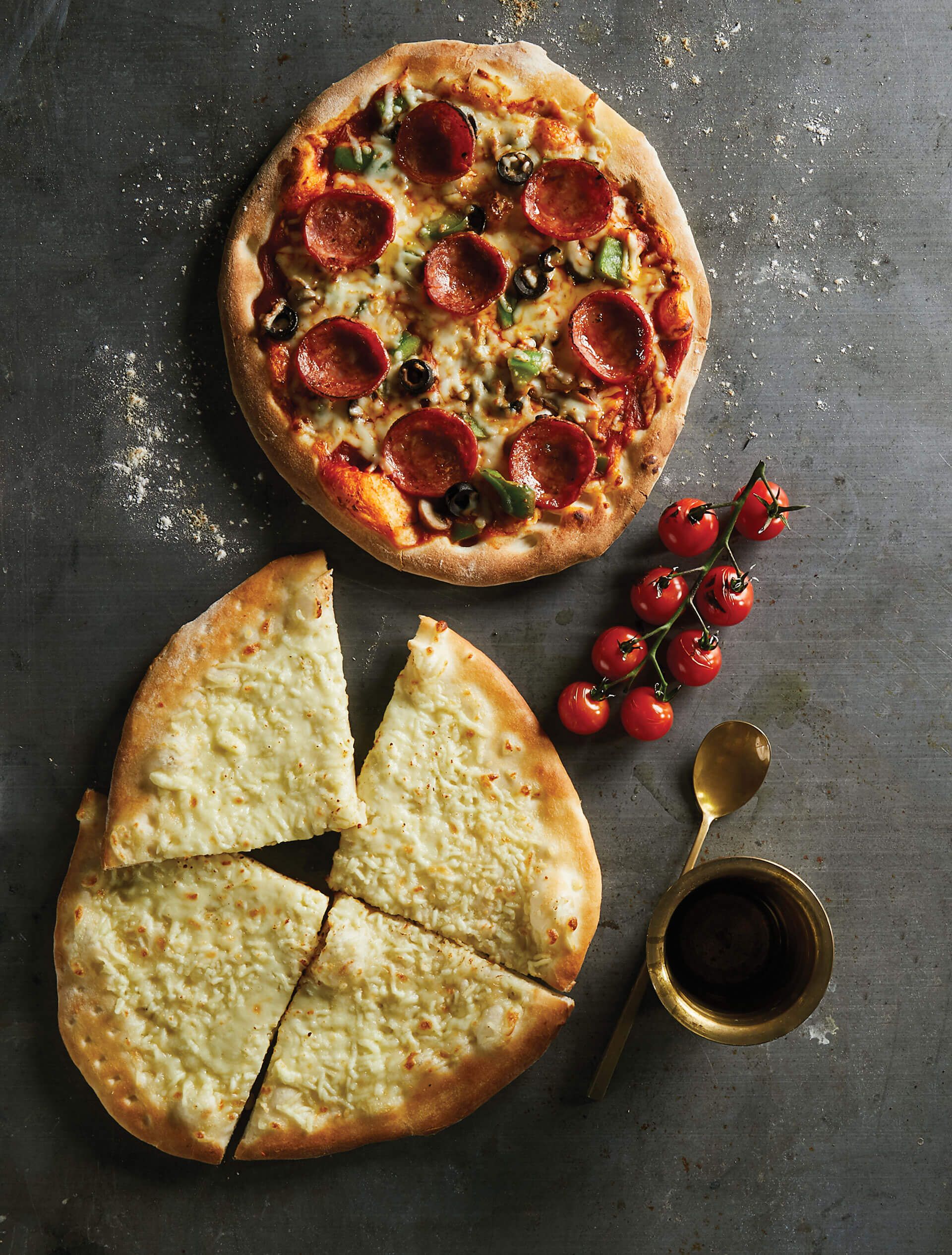 Two pizzas are on a table with tomatoes and a spoon