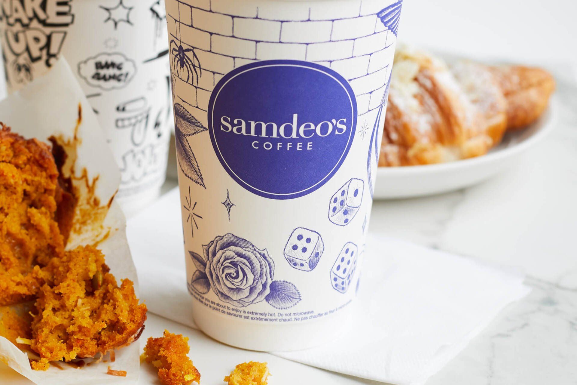 A cup of samdeos coffee is sitting on a table next to a plate of food.