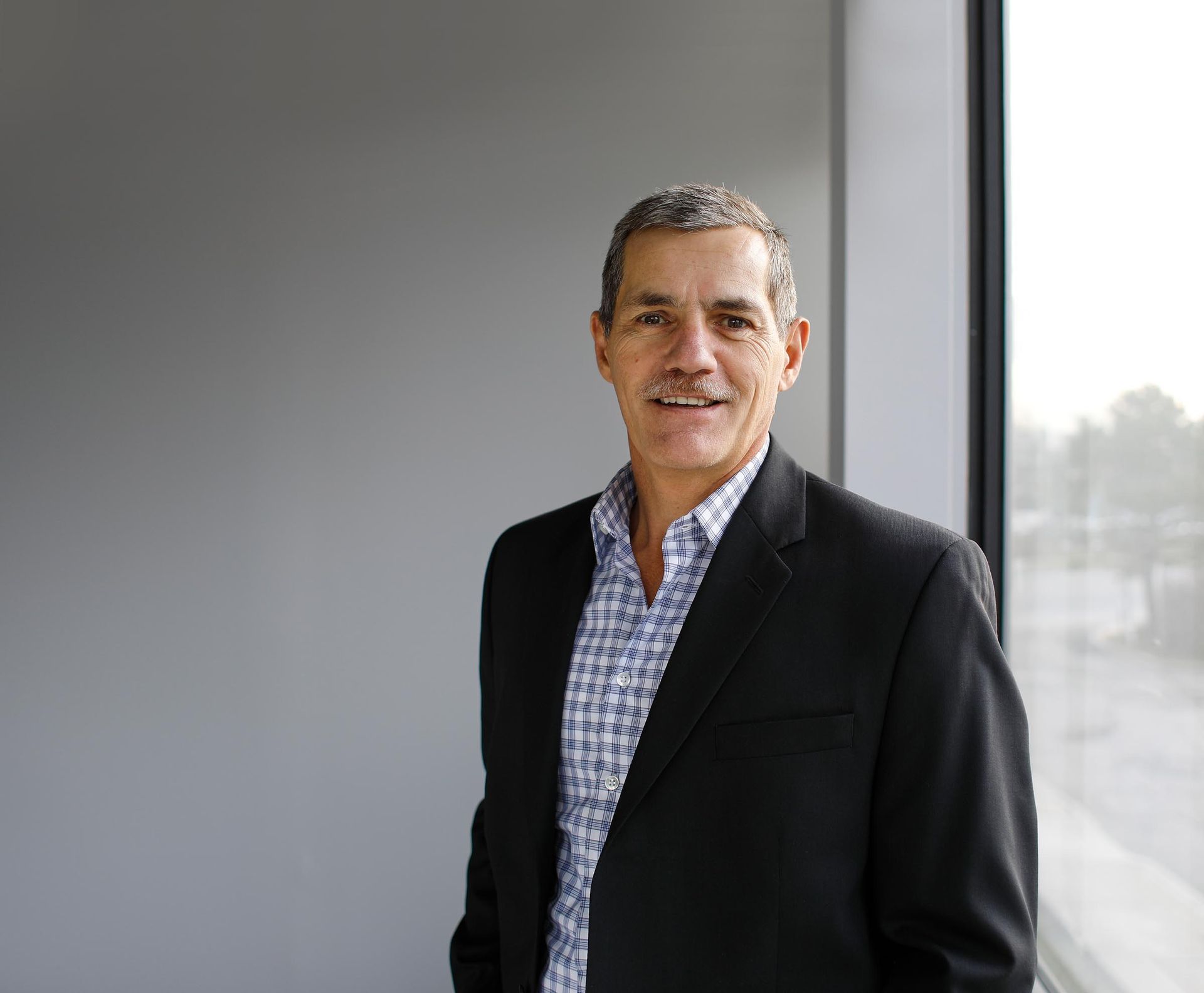 A man in a suit and plaid shirt is standing in front of a window.